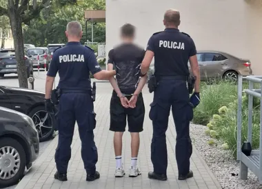Dwóch policjantów w mundurach z napisem POLICJA prowadzi zatrzymanego mężczyznę w kajdankach chodnikiem przy budynku i zaparkowanych samochodach.